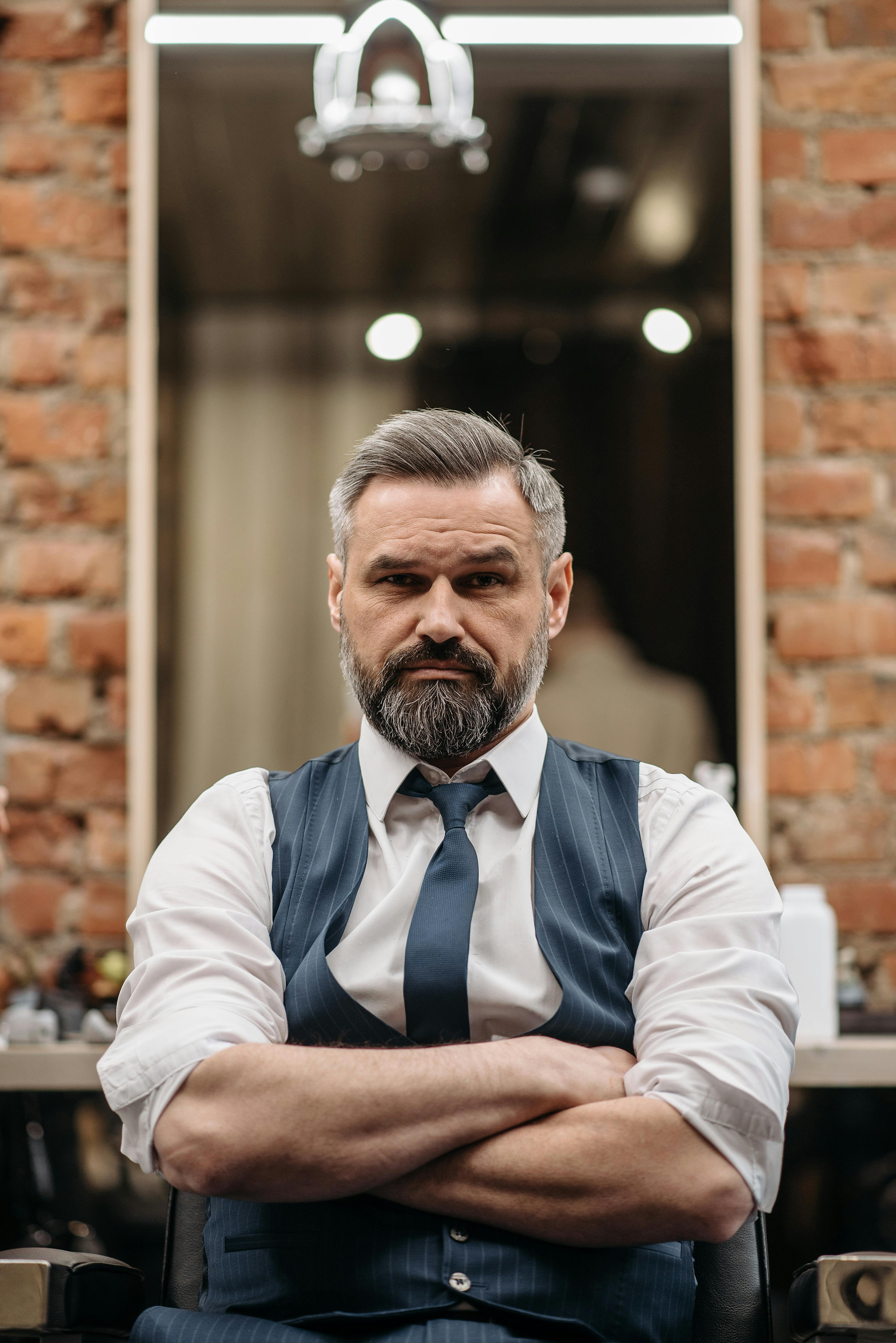 Middle-aged man with a beard, arms crossed. He is wearing a blue vest, white shirt and a tie. Barbershop interior.