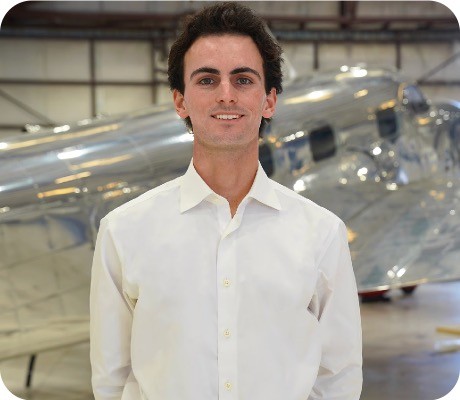 A young man in a white shirt stands in front of an airplane in a hangar, smiling at the camera.