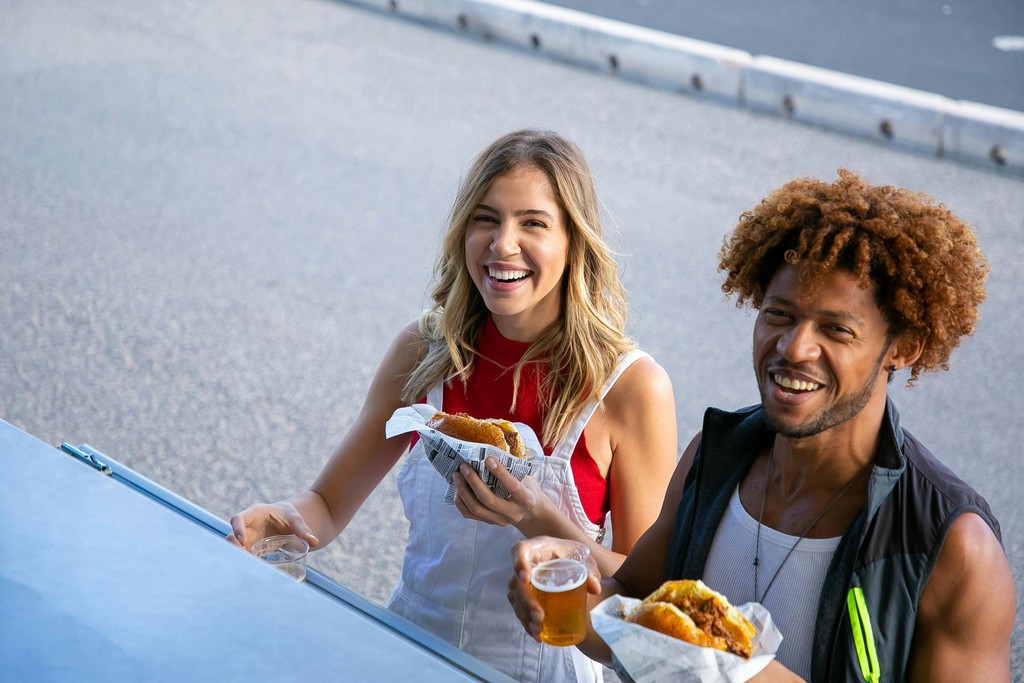 Two smiling young adults holding fast food burgers and drinks outside a food vendor, representing newcomers enjoying budget-friendly takeout meals in Canada.