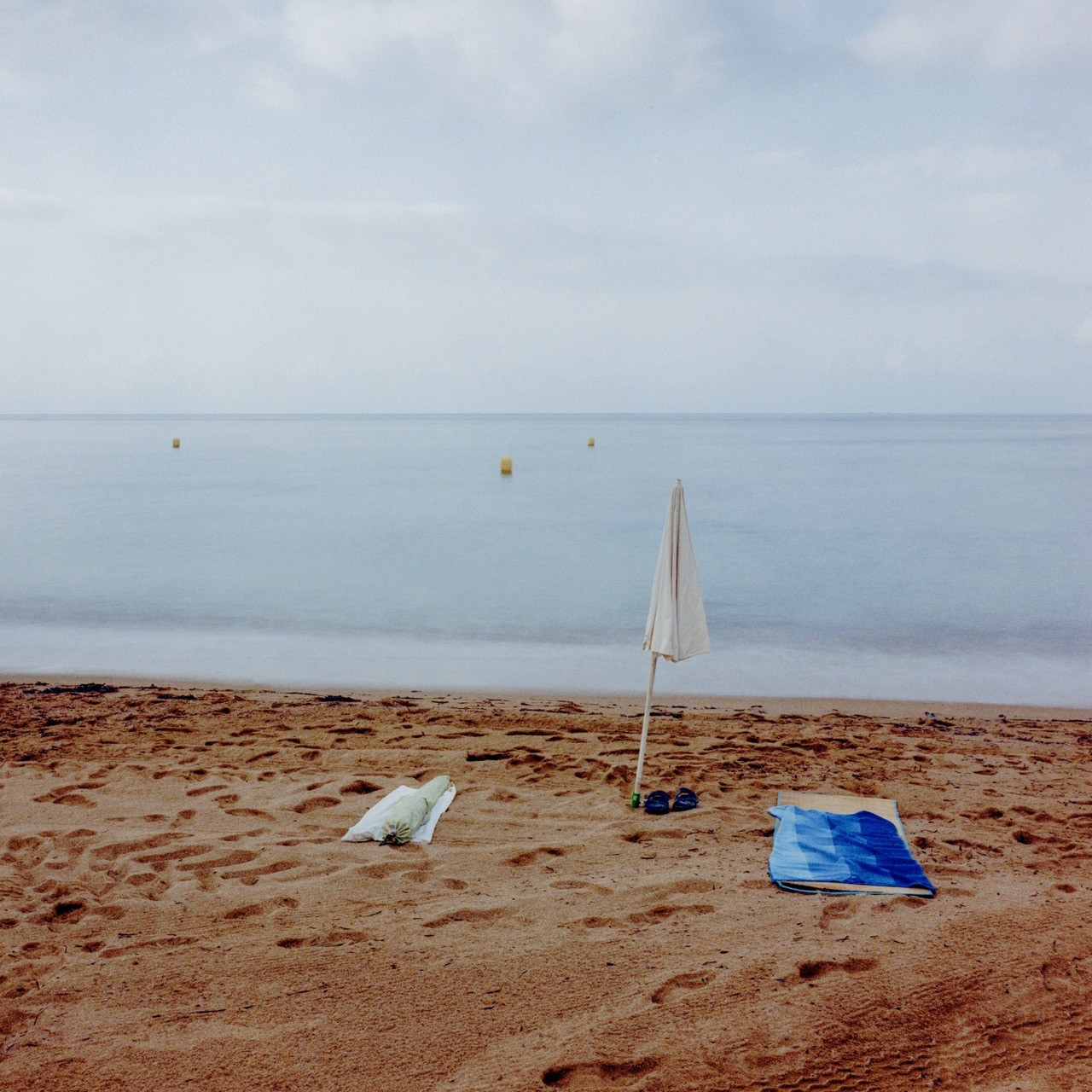 A closed parasol, sandals and a folded blue towel rest on an empty beach, The Side of Summer by Renzo Cicillini, analog medium-format fine art photography.
