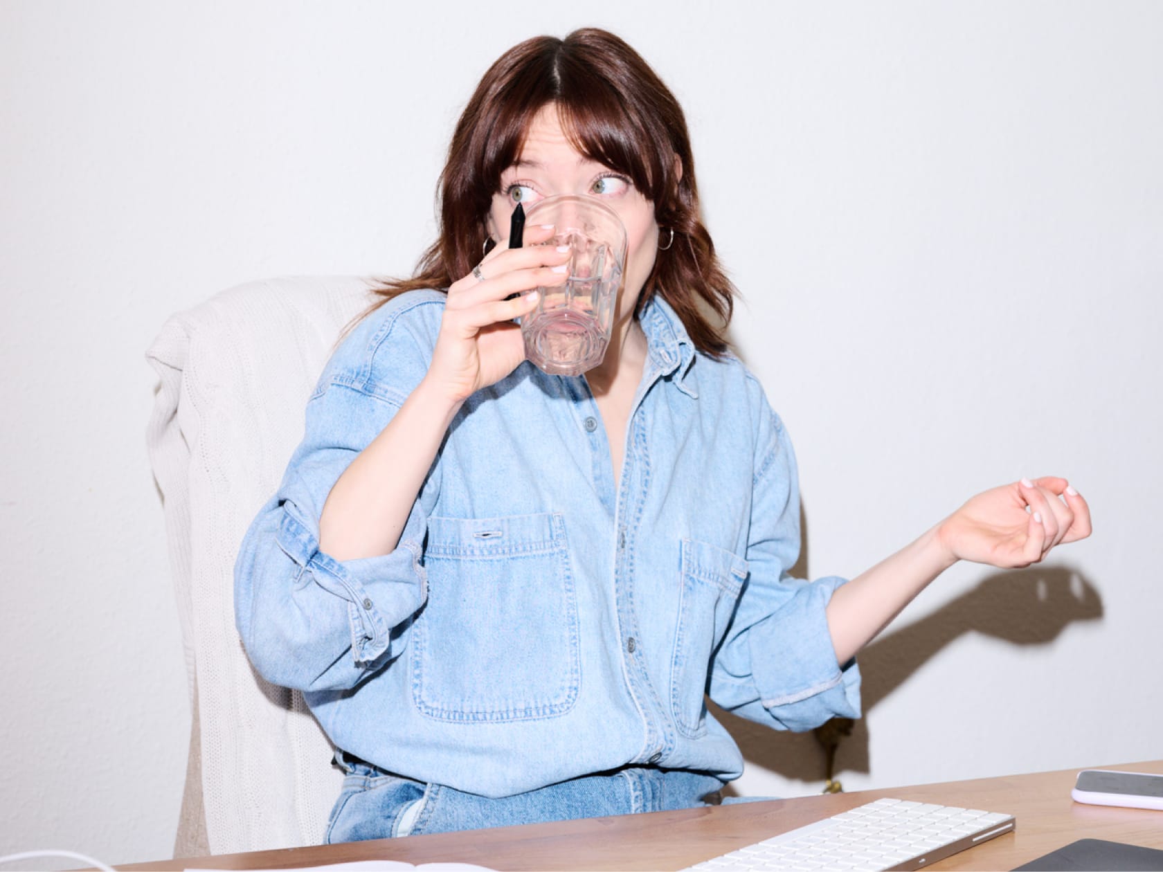 Woman in a denim shirt drinking water while sitting at a desk with a keyboard and phone