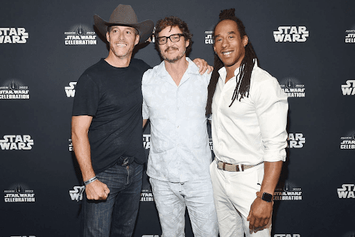 Three men smile together at a red carpet event, dressed in stylish outfits against a dark backdrop.