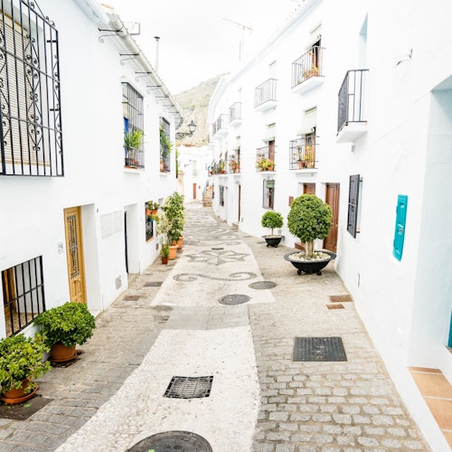 En smal, kullerstensbelagd gata kantad av vita byggnader, krukväxter och smidesjärnräcken, som leder uppför en svag backe.