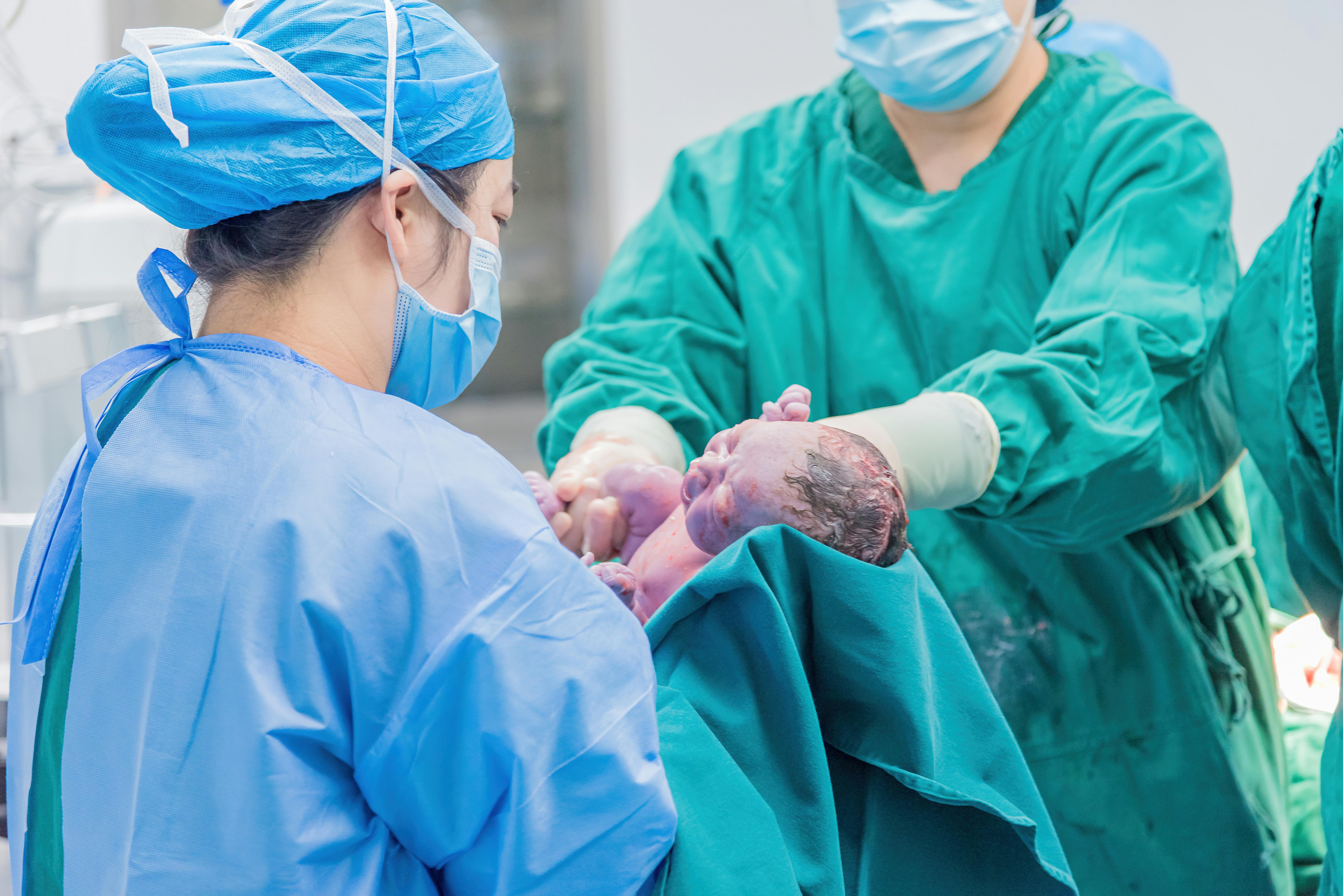 A surgeon is placing a newborn baby delivered through caesarean section onto a turquoise medical cloth held by another medical practioner in blue scrubs.