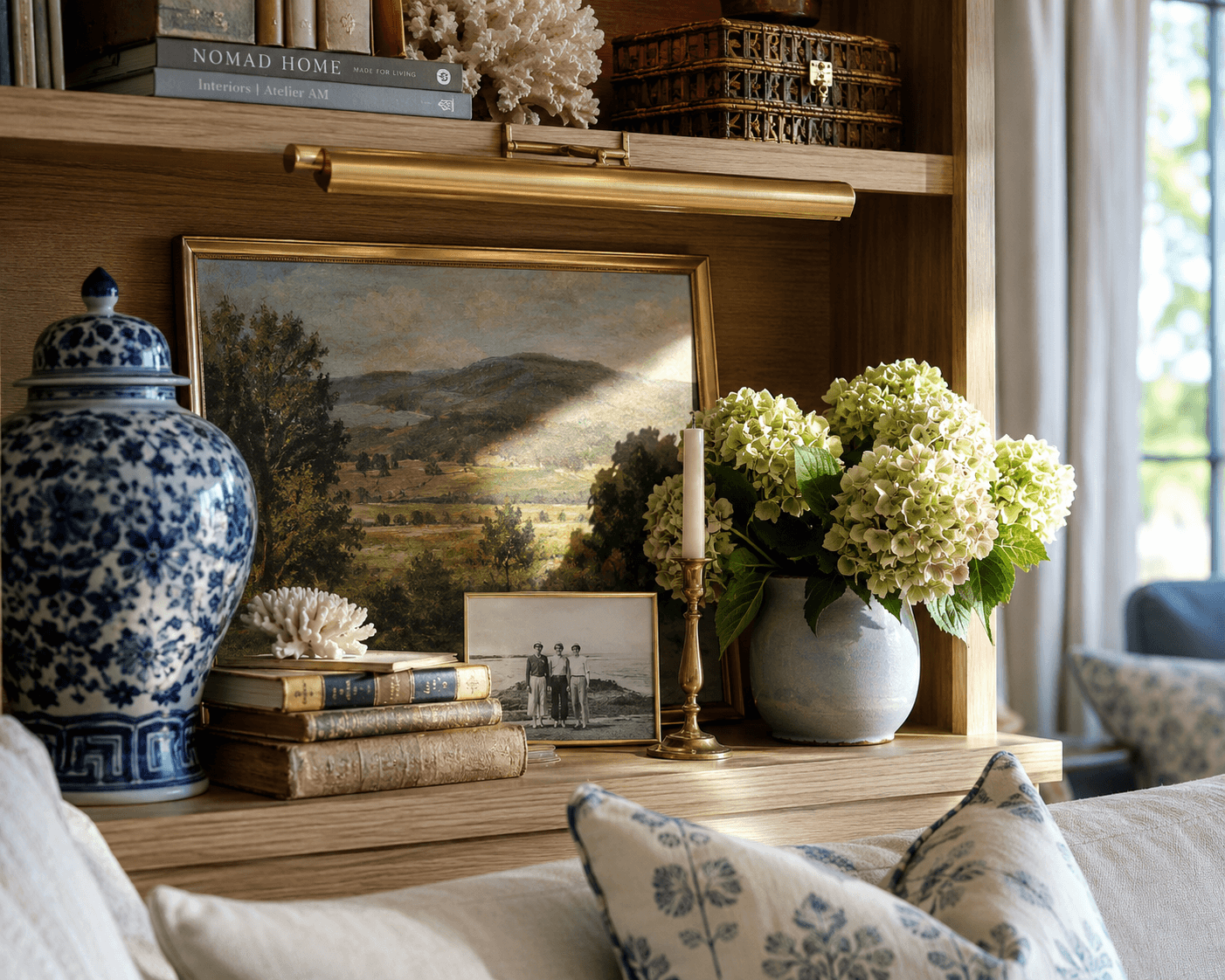 Elegant living room bookshelf styled with blue and white ceramic vase, vintage books, framed landscape painting, brass candle holder, fresh hydrangea flowers in a ceramic pot, and decorative coral, creating a cozy and sophisticated home interior.