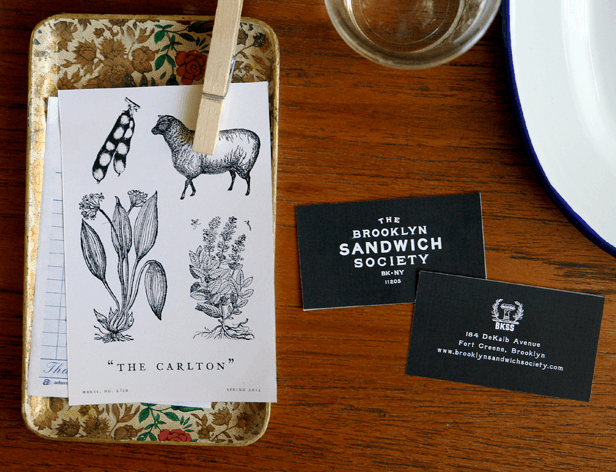 The image shows a wooden table with a decorative tray holding two business cards and a printed sheet of paper. The paper sheet displays a black and white illustration labeled “THE CARLTON,” depicting a sheep and a plant with leaves and flowers. A wooden clothespin secures the printed sheet to a clipboard on the tray. The tray rests on a wooden table surface. To the right of the tray, a clear glass or cup is visible, and a white plate is partially seen. The business cards are black with white text and display the names “THE BROOKLYN SANDWICH SOCIETY” and “BK-NY 11805,” along with a website address: www.brooklynsandwichsociety.com.