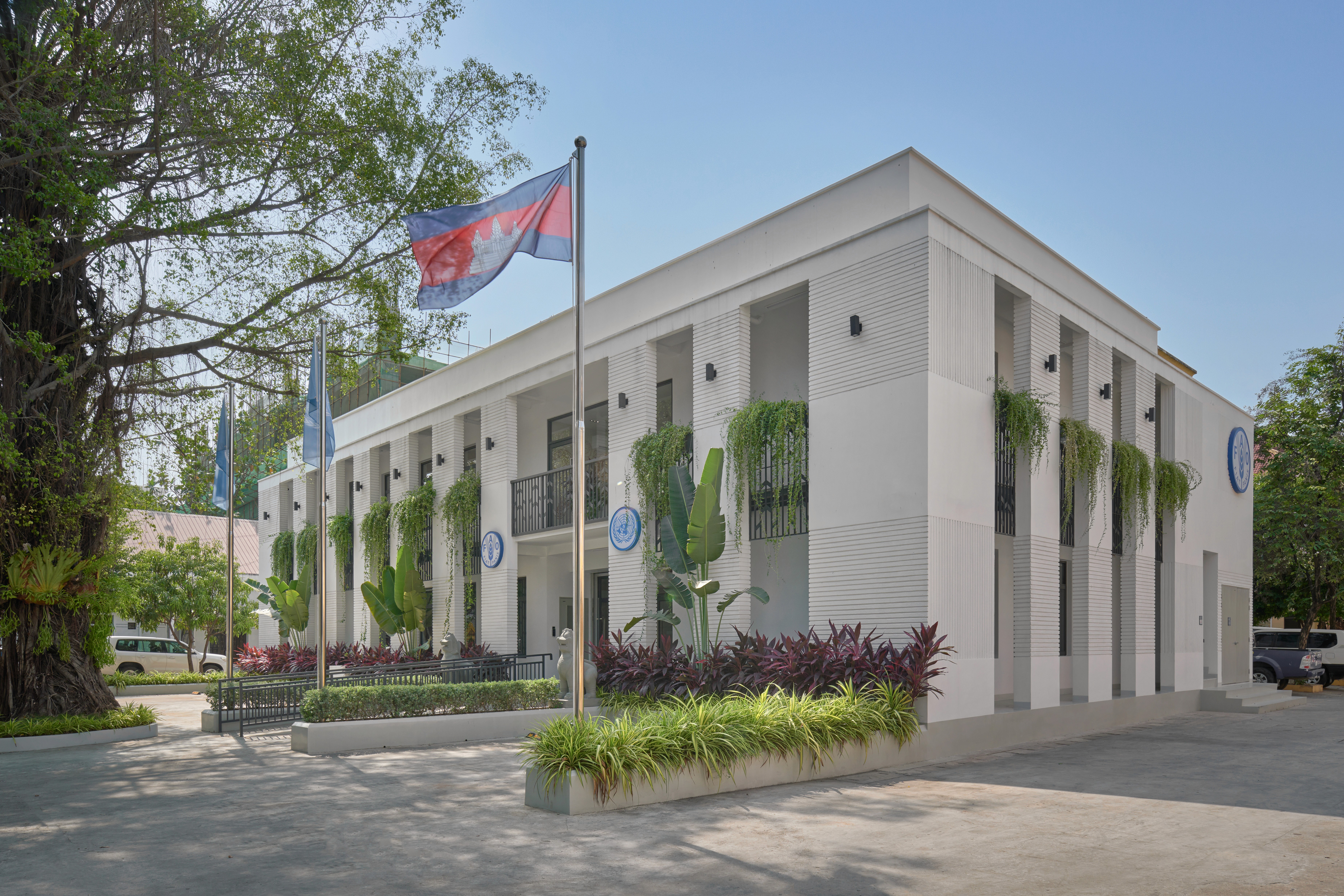 Modern white two-story office building exterior with vertical architectural fins, lush tropical landscaping, and the Cambodian flag at the entrance.