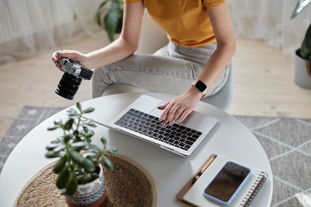 Photographer working on laptop while holding camera at home