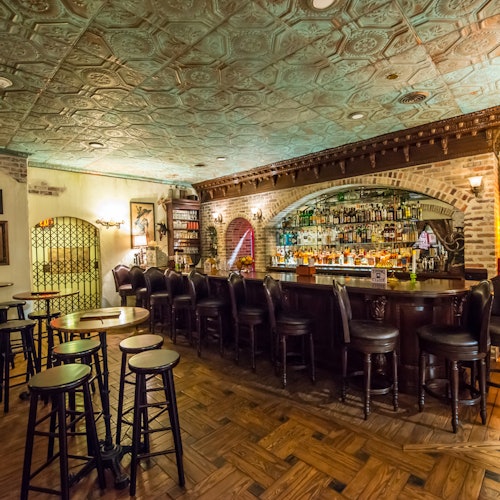 一个温馨且灯光柔和的酒吧，拥有复古装饰，特色为木质吧台、带软垫的吧凳和摆满酒瓶的架子。