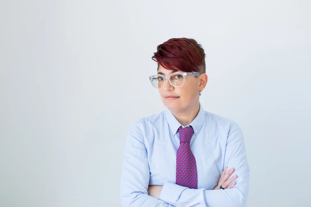 A woman with short, dark red hair and transparent glasses is wearing a light blue dress shirt and a red patterned tie, standing against a plain white background with arms crossed in a confident posture.