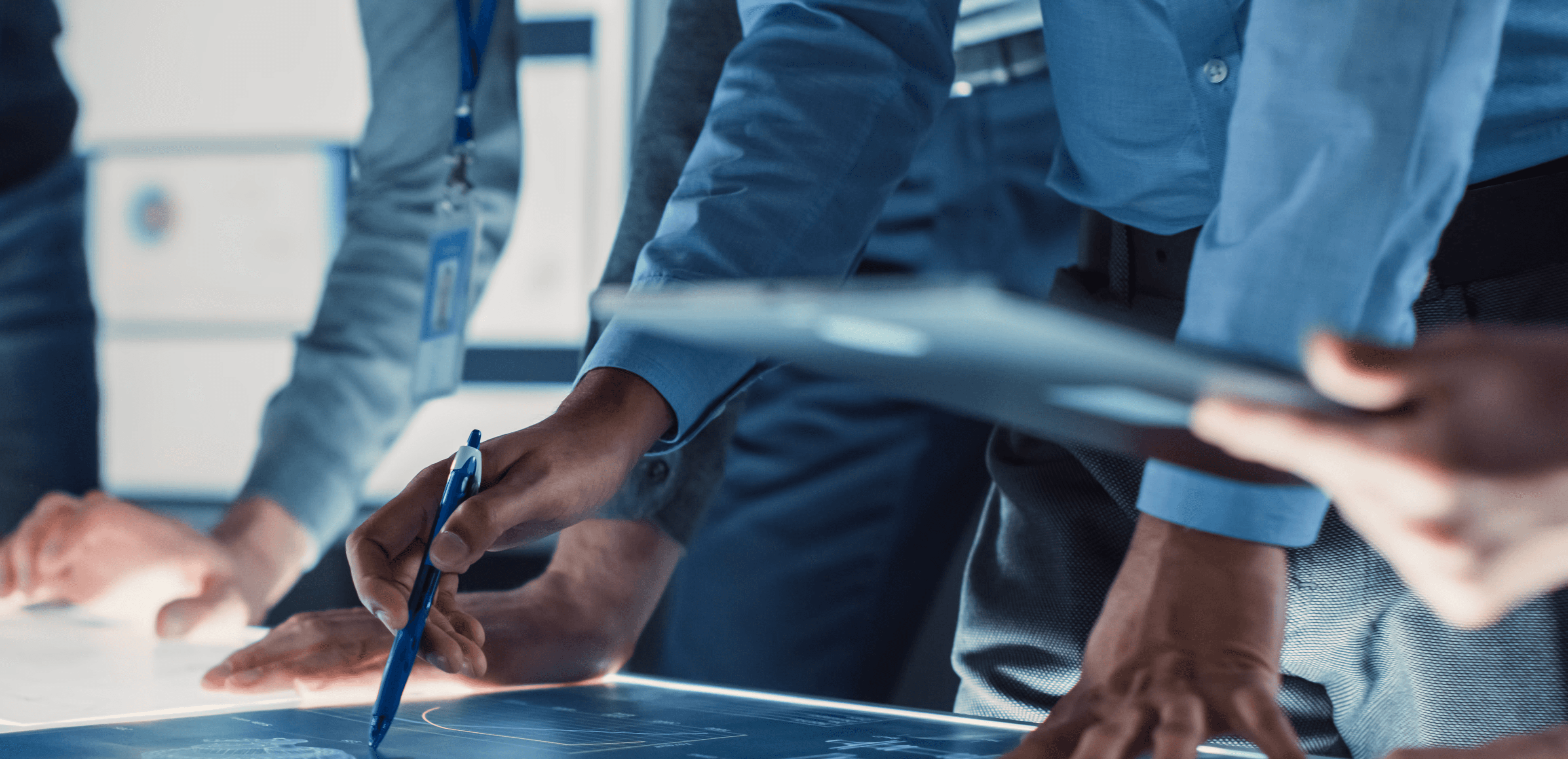 A diverse team of professionals collaborating around an illuminated digital display table, reviewing data and plans in a modern work environment.