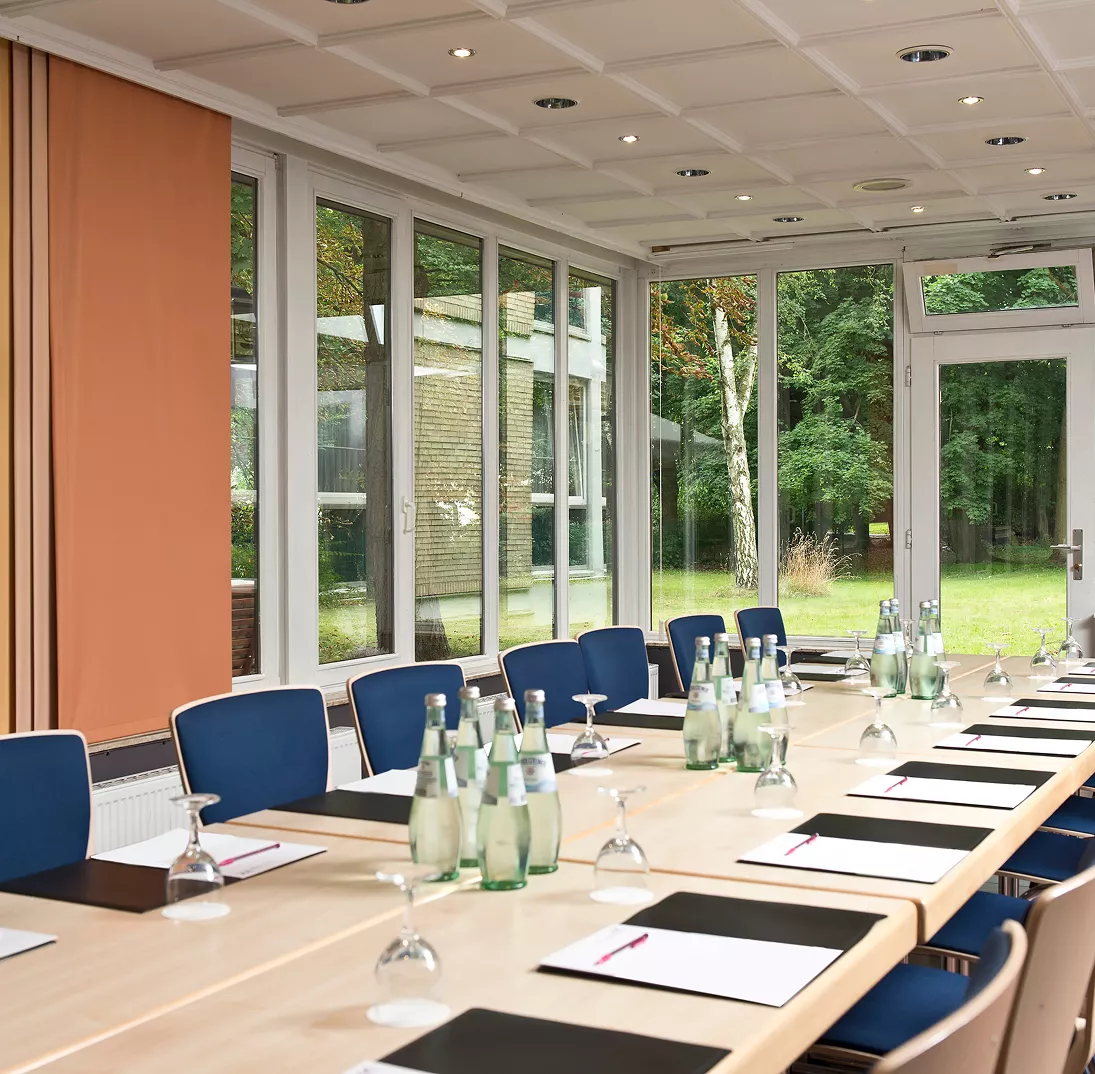 Aufnahme eines Konferenztisches mit blauen Stühlen. Der Raum verfügt über Fenstern, die ins Grüne schauen