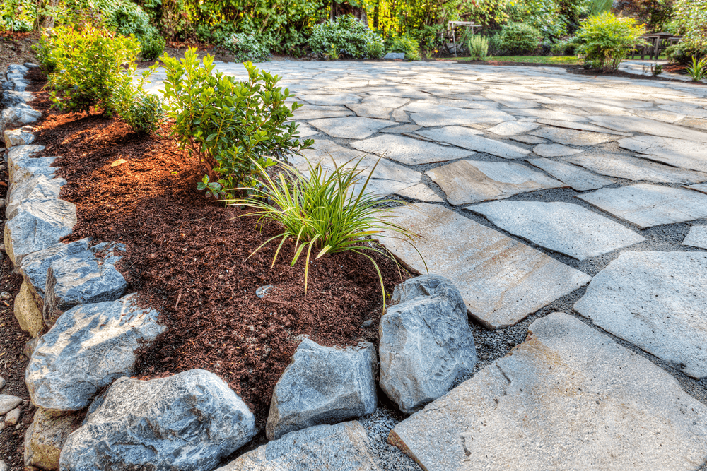 A natural stone patio bordered by mulch and rocks, featuring small bushes and ornamental grasses. The earthy tones and irregular flagstone shapes create a rustic, organic outdoor space.