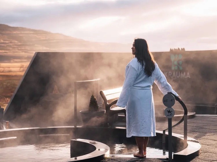 Kona í hvítum baðsloppi stendur við gufandi jarðhitalaug á útispa og horfir út yfir rullandi hæðir undir móðuðu sólsetri. Krauma-merkið sést á vegg í bakgrunni.