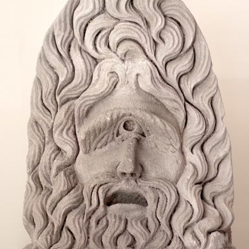 Một bức tượng điêu khắc bằng đá của một khuôn mặt có râu với một con mắt ở giữa trán, được bao quanh bởi mái tóc xoăn.