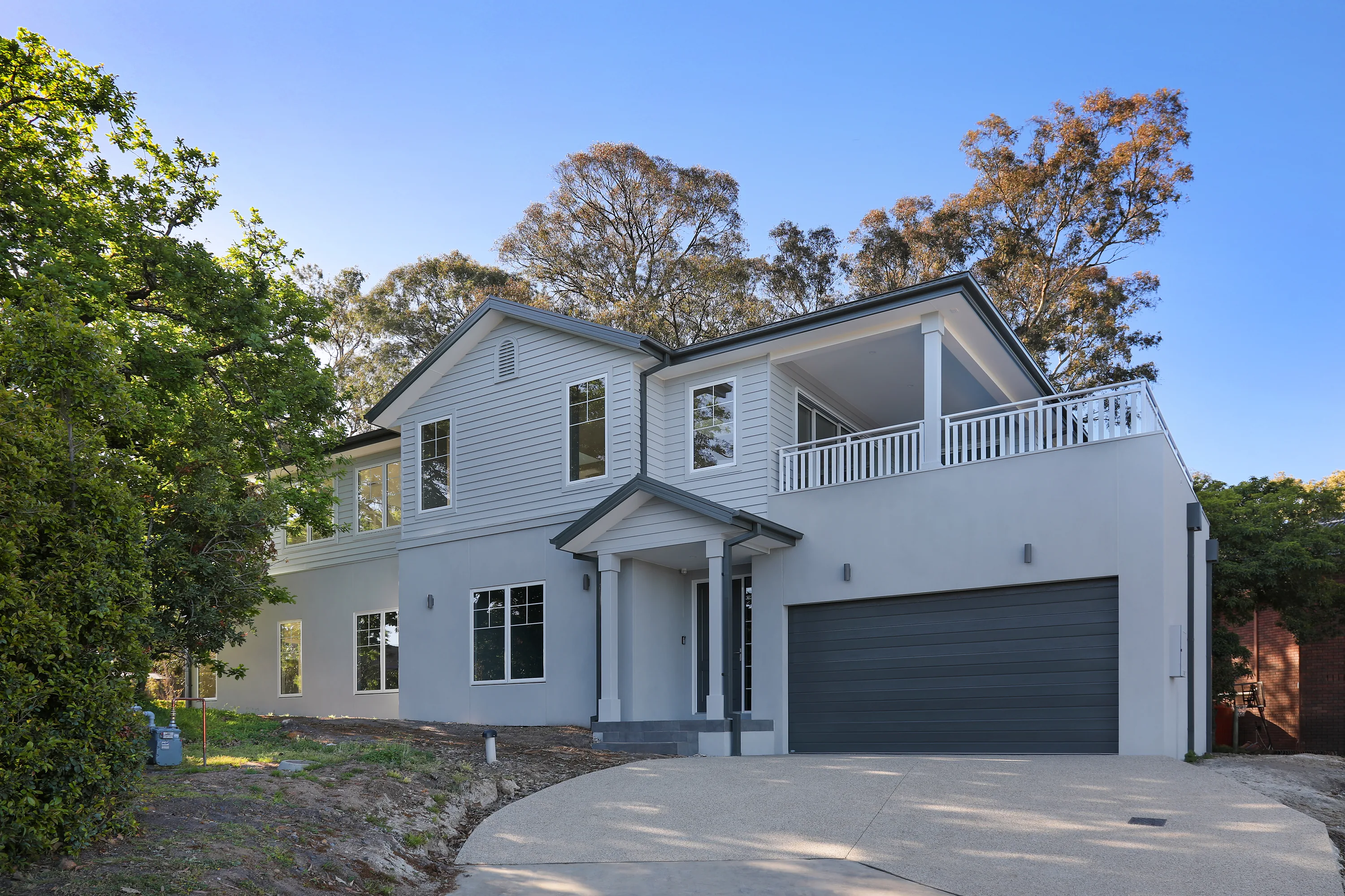Front of the Eltham Residence among gum trees, a two-storey pale weatherboard and rendered home with white-trimmed windows, gabled entry portico, columned balcony and a charcoal double garage off a curved driveway