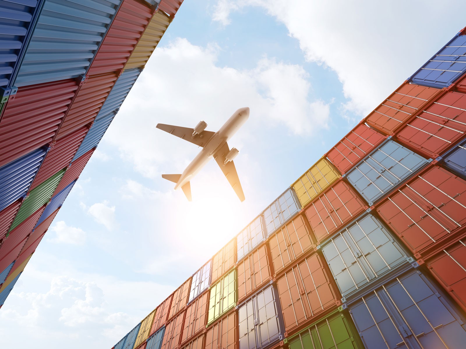 A cargo airplane flying overhead with a view from below framed by stacked colorful shipping containers, symbolizing global logistics and transportation.