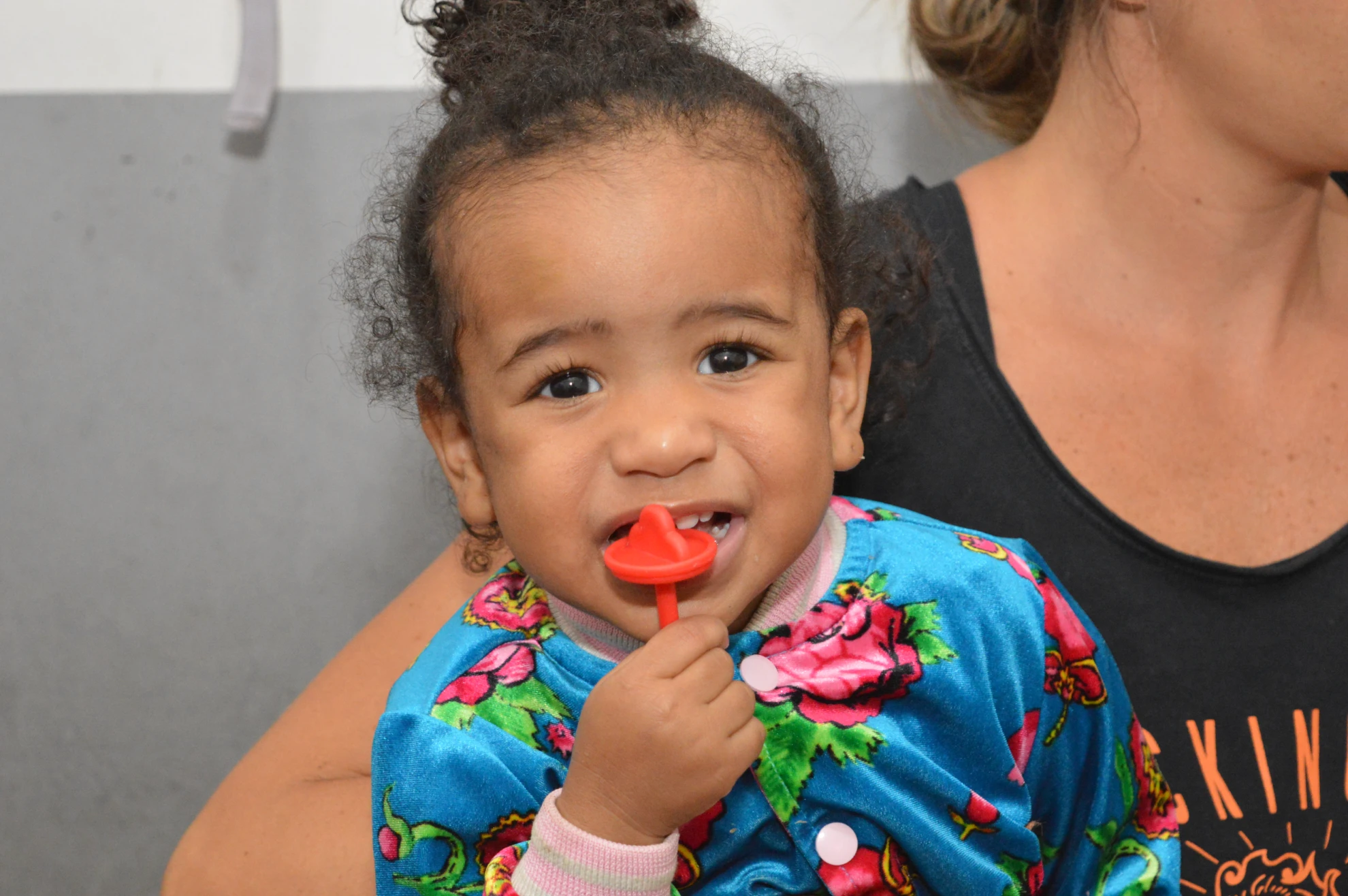Menina com vestido colorido no colo de uma adulta, olhando para a câmera com expressão curiosa.