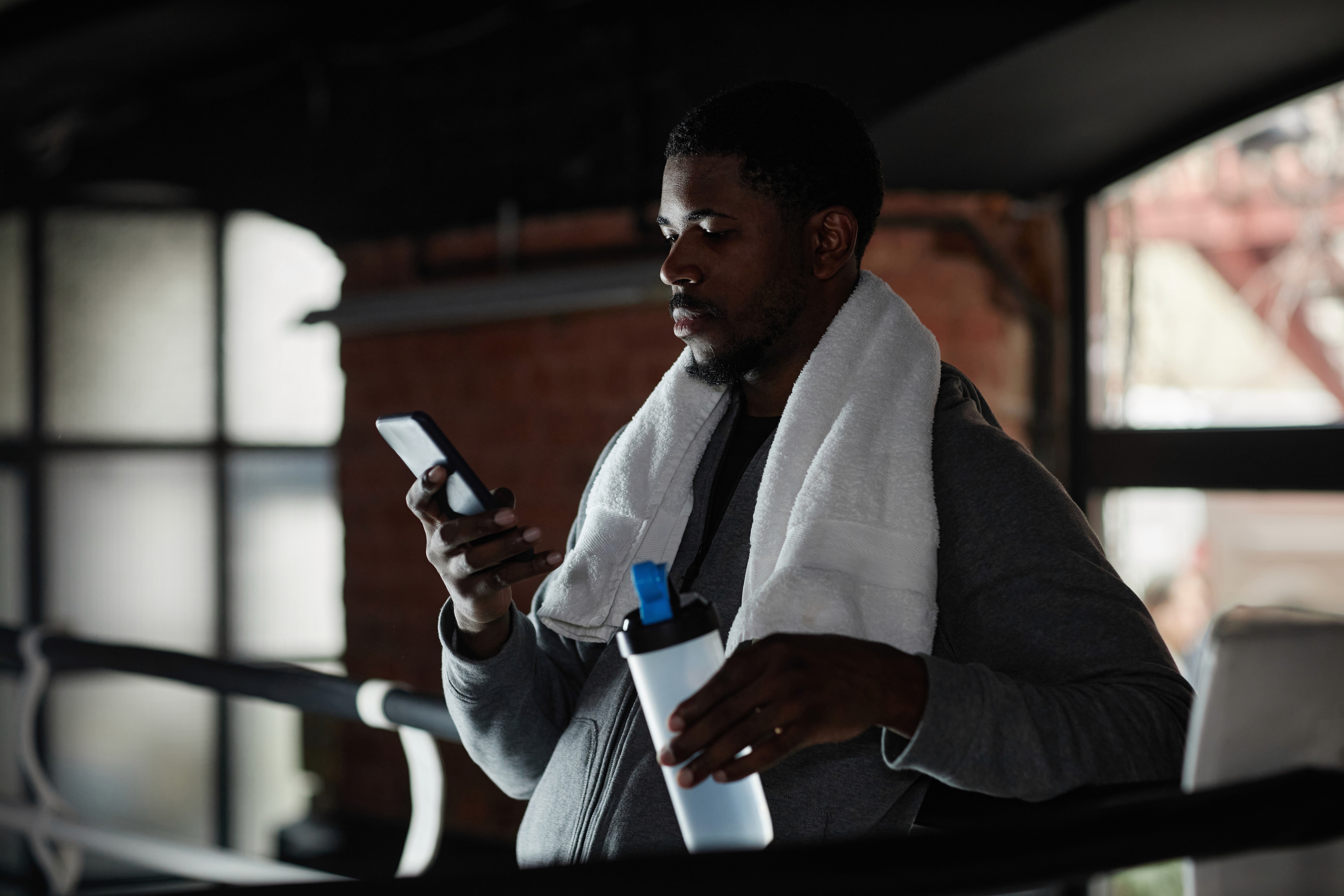 A young man standing in a gym with a white towel draped over his shoulders, holding a shaker bottle in one hand while looking at his smartphone with the other. He is wearing a gray hoodie, and the scene is lit softly, creating a relaxed atmosphere. The background includes windows and a brick wall, providing a casual, indoor workout setting. The image conveys a moment of rest or focus between exercises, as he checks his phone, possibly tracking his workout progress or taking a break.