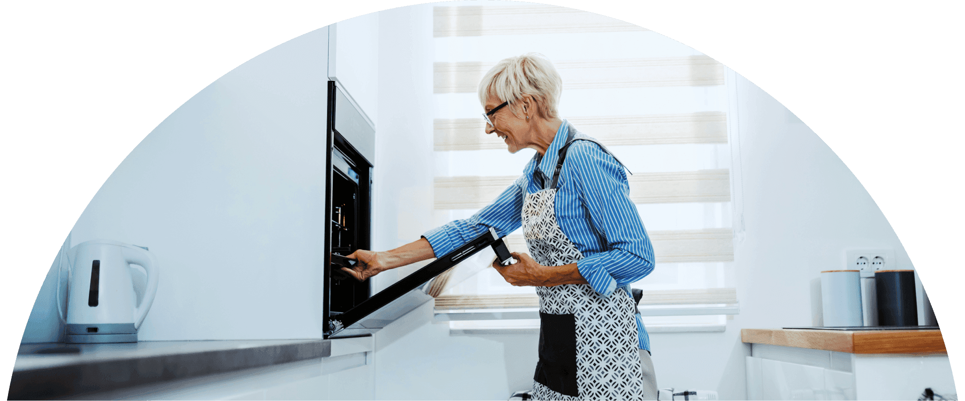 Older woman wearing an apron places a dish into a built in oven in a bright modern kitchen