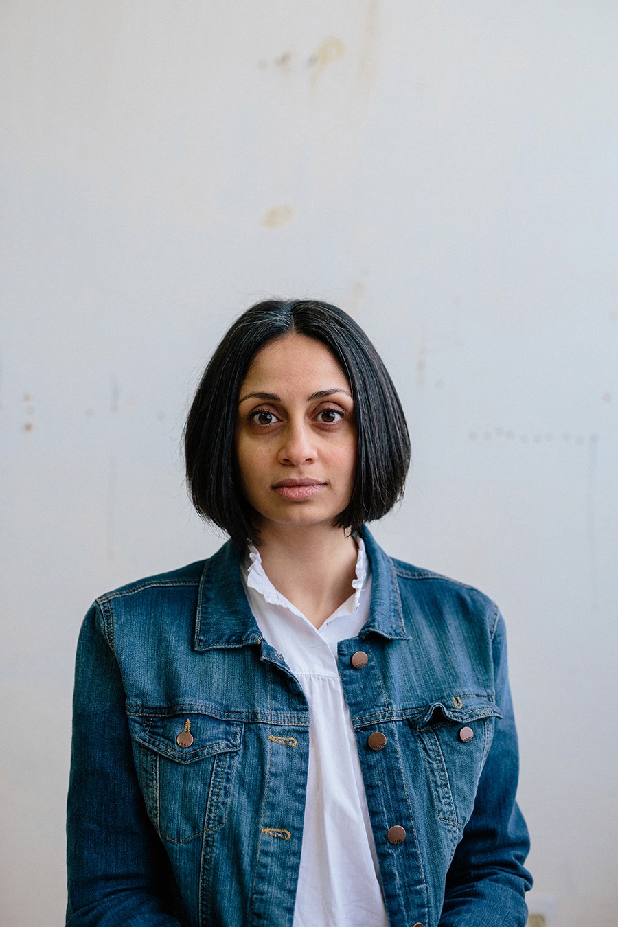 A person with a bob haircut wearing a denim jacket stands against a plain white wall, looking directly at the camera.