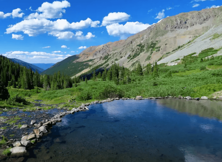Rock-edged pool overlooking a green mountain valley beneath steep rocky ridges and a blue sky