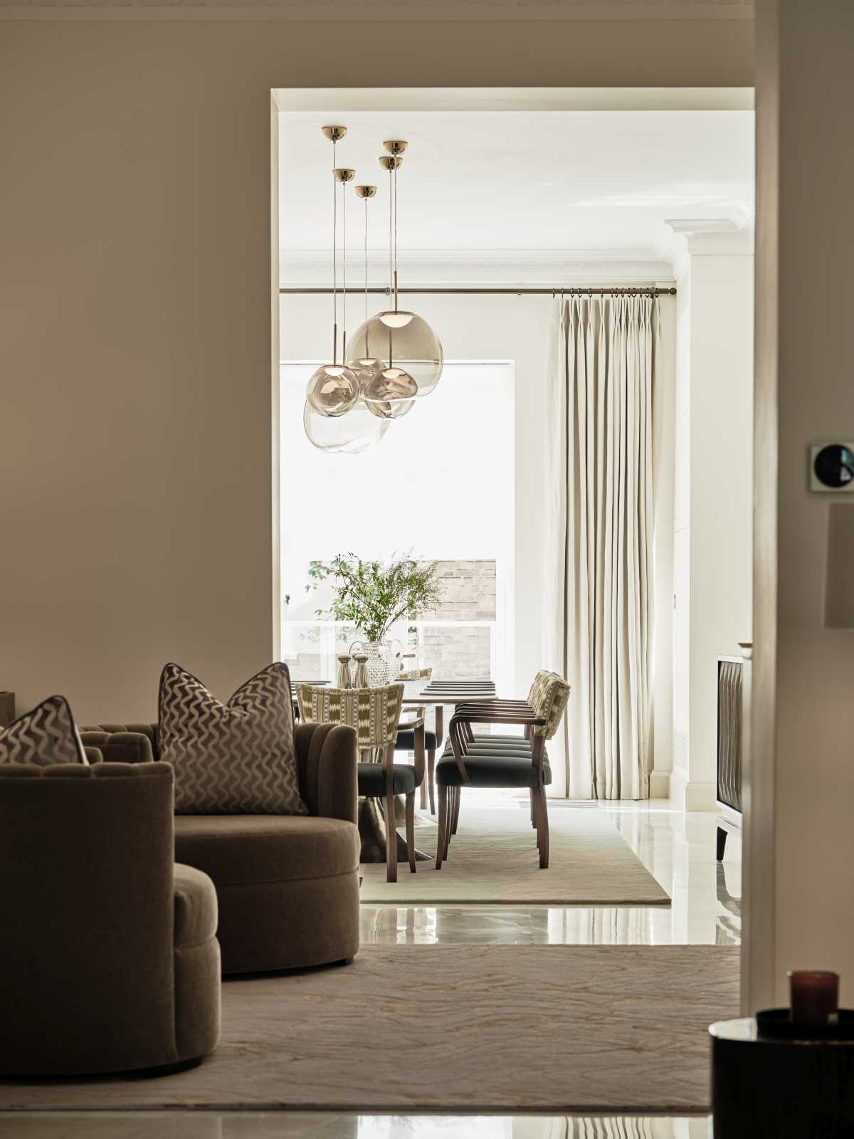 Looking through from a sophisticated sitting area into a brightly lit dining room. The foreground features plush, low-profile taupe armchairs on a neutral rug, capturing the elegant ceremonial flow and spatial planning of a premium central London residence.