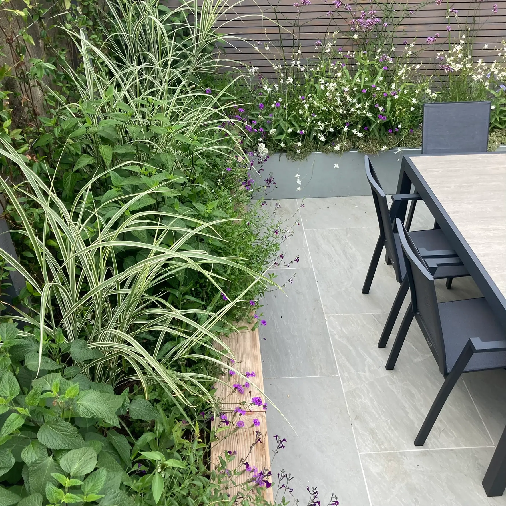 A garden scene featuring lush greenery beside a stone pathway and a black outdoor dining table with chairs.