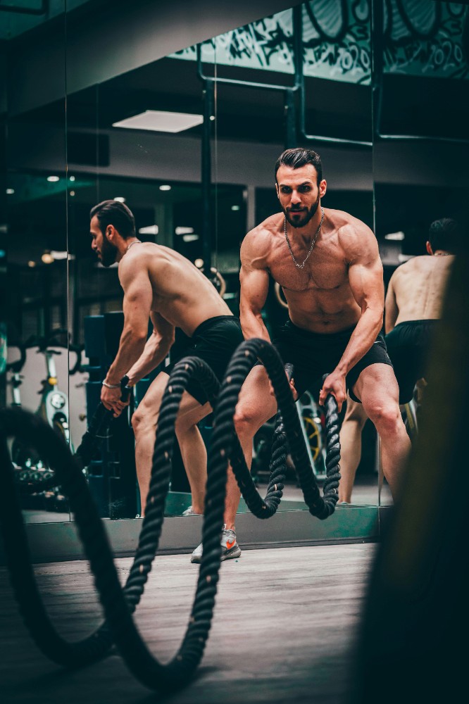 A shirtless man intensely using battle ropes in a gym, with another man in the background.