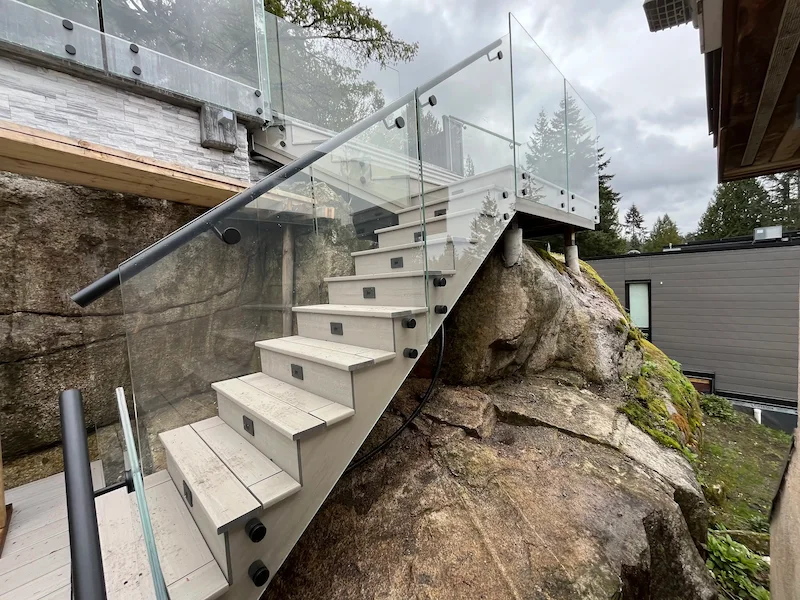 Exterior glass stair railing on an outdoor staircase with side-mounted glass panels and metal handrail built into a rock landscape.