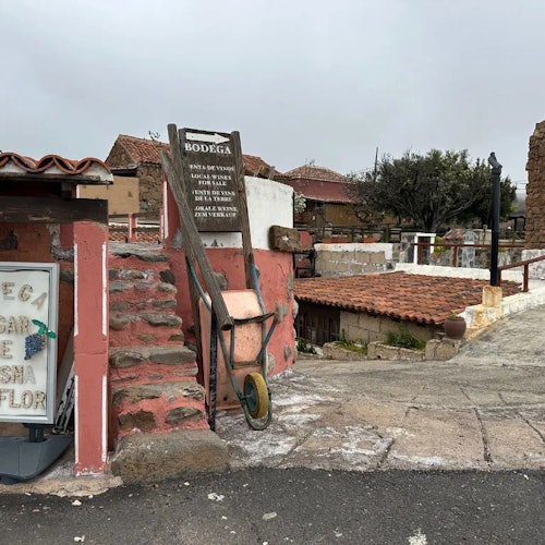 乡村场景，砖墙、旧手推车、石阶及一家小酒馆的招牌。背景是瓦屋顶和多云的天空。