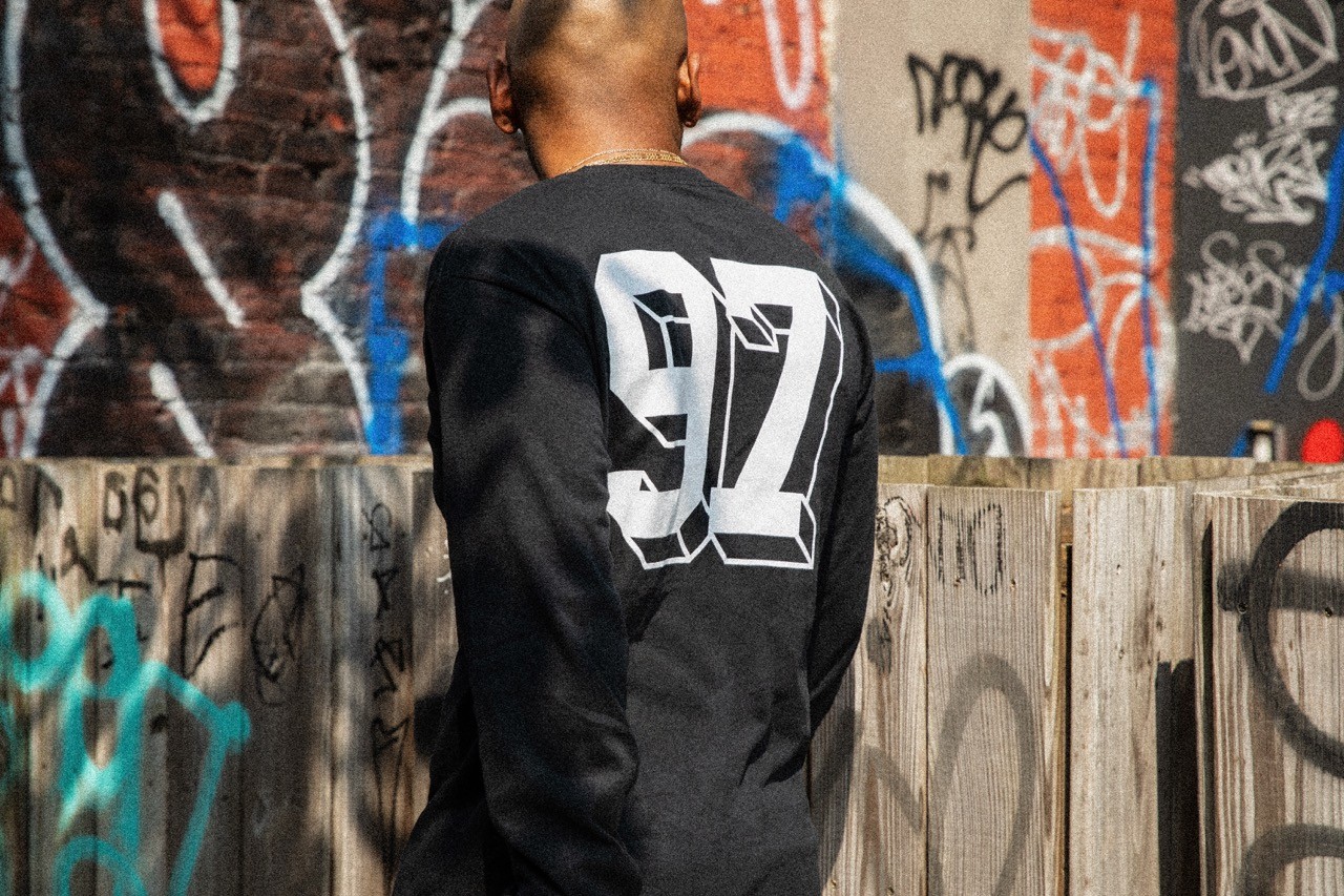 Back view of model wearing a black top with a bold ‘92’ graphic, photographed against a brick wall.