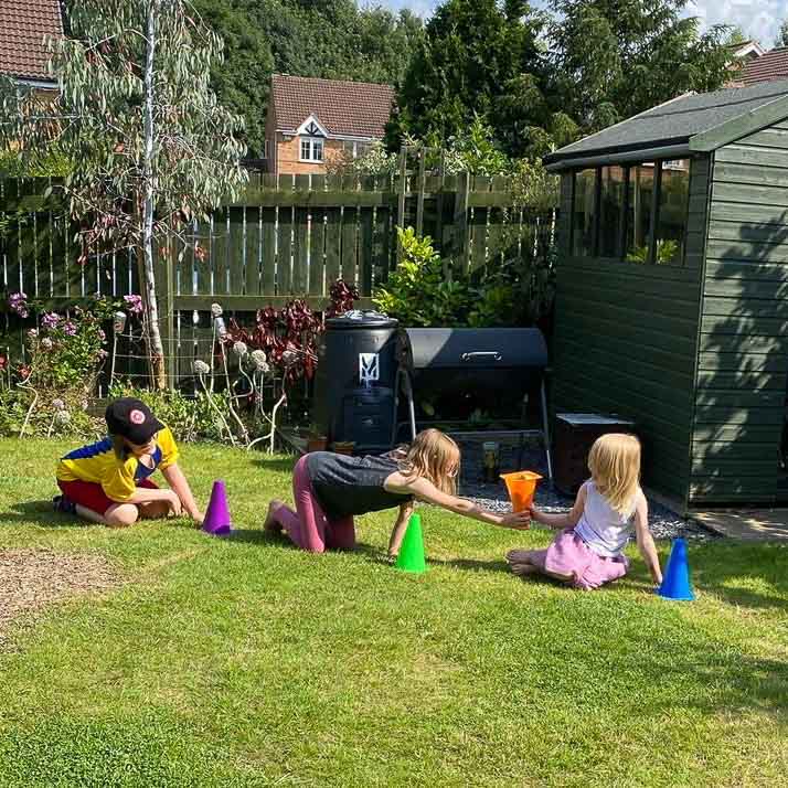 Three children are sat in a sunny garden between a line of small cones. They are playing a game where they are passing objects inside one of the upturned cones.