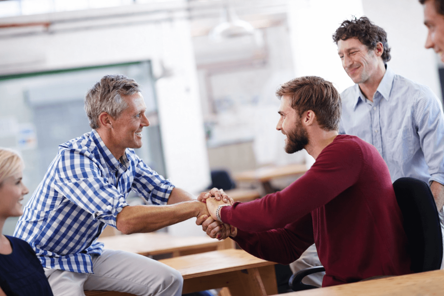 Two colleagues smiling while working together at a desk, one wearing a cream jacket and the other a beige cap and glasses.