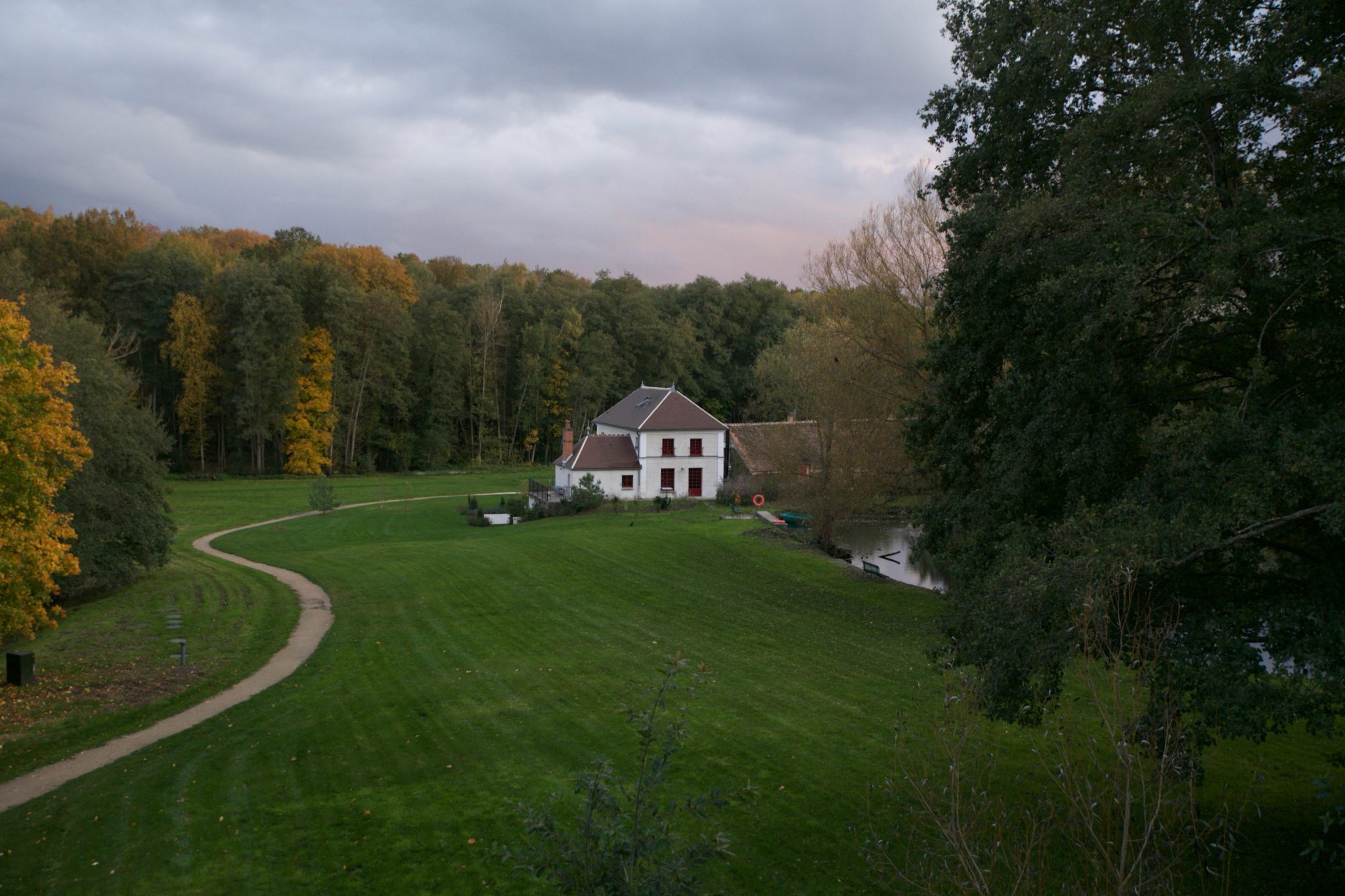 White manor house beside a pond at dusk, seen across a sweeping lawn and curved path