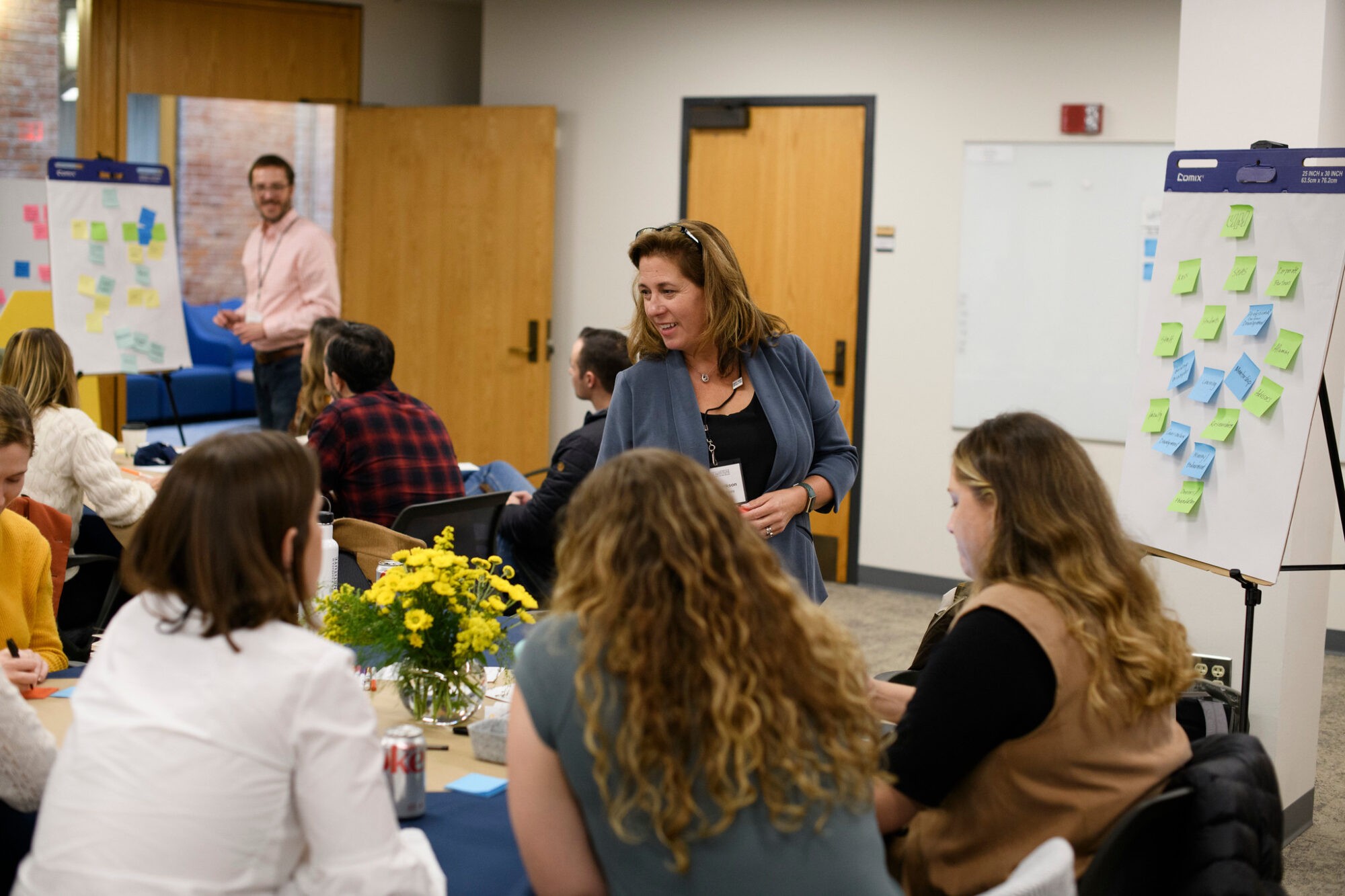 A woman speaks to a group seated at tables, engaged in discussion, with colorful flower arrangements present.