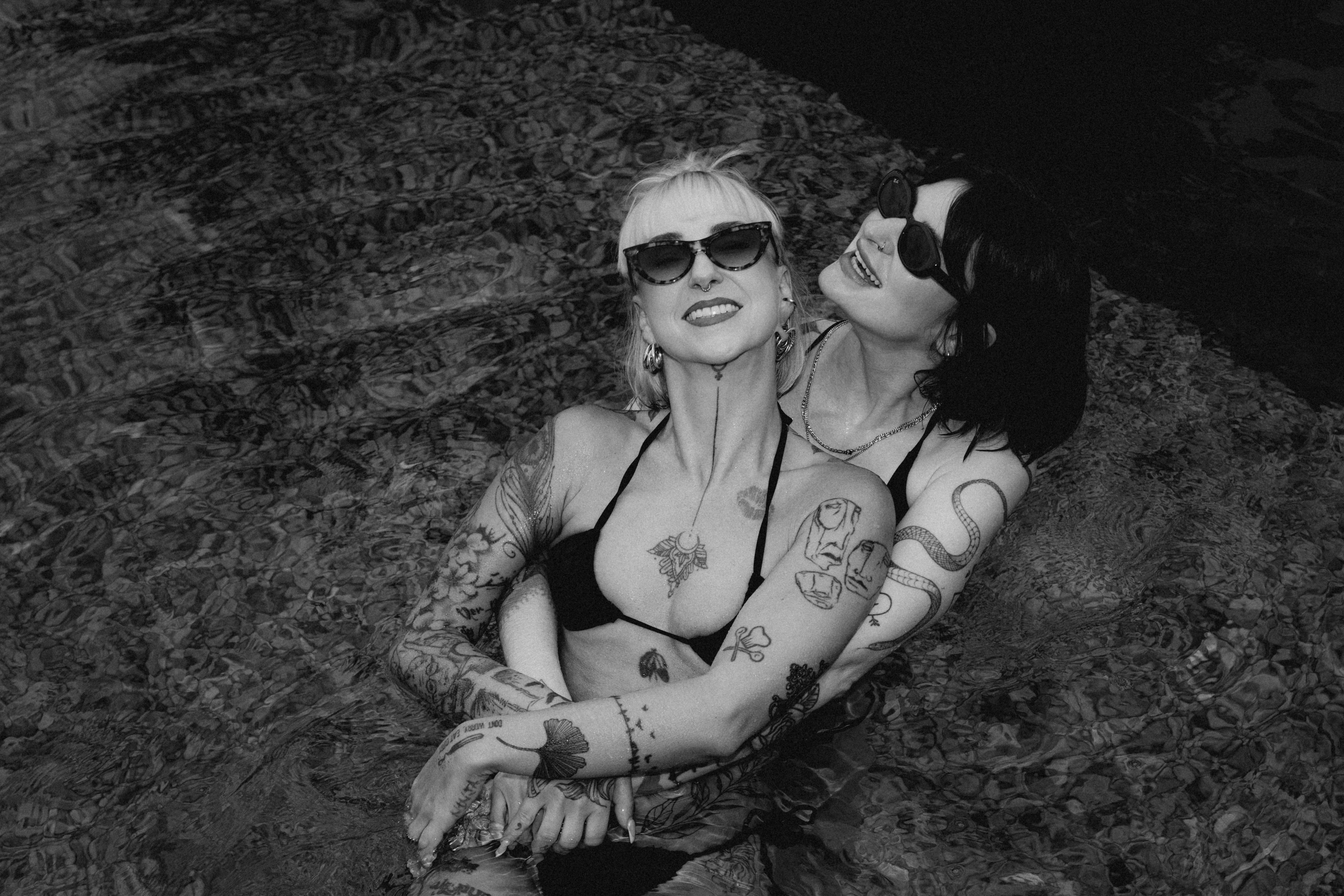 Two women in swimwear relax together, wearing sunglasses, against a dark background, captured in black and white.