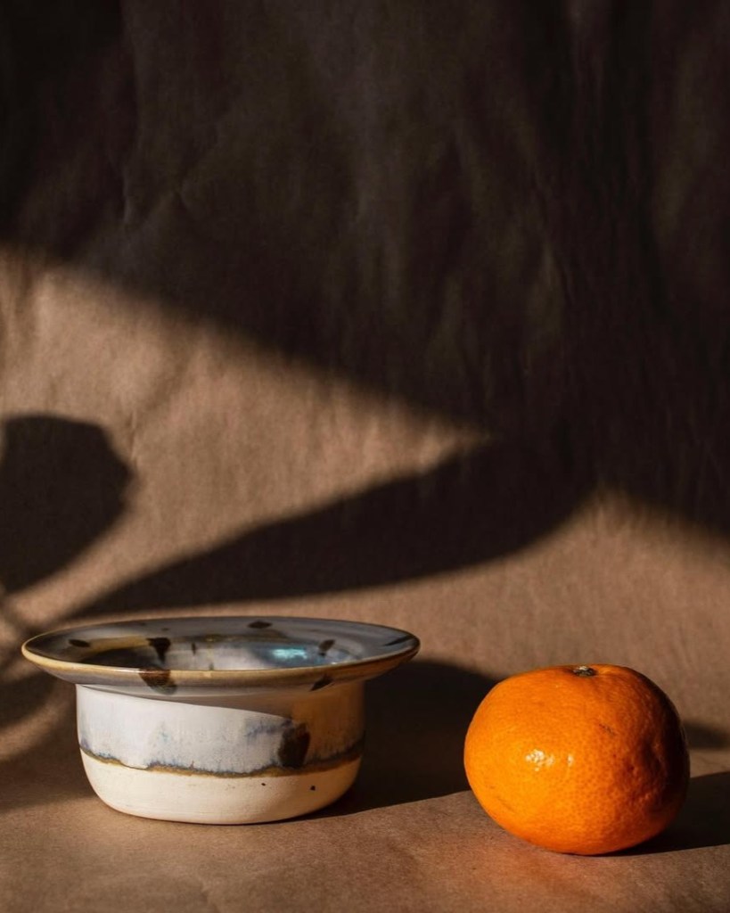 A ceramic bowl with a wide rim and decorative patterns sits next to a bright orange fruit on a textured brown surface.