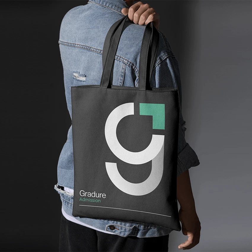A charcoal tote bag with a large white "g" monogram, mint corner mark and "Gradure Admission" text, held over the shoulder of a person in a denim jacket.