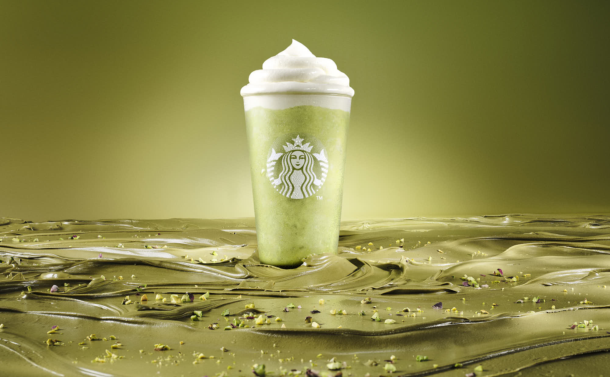 A tall, illuminated pistachio coffee stands against a greenish backdrop, surrounded by a pistachio cream river.