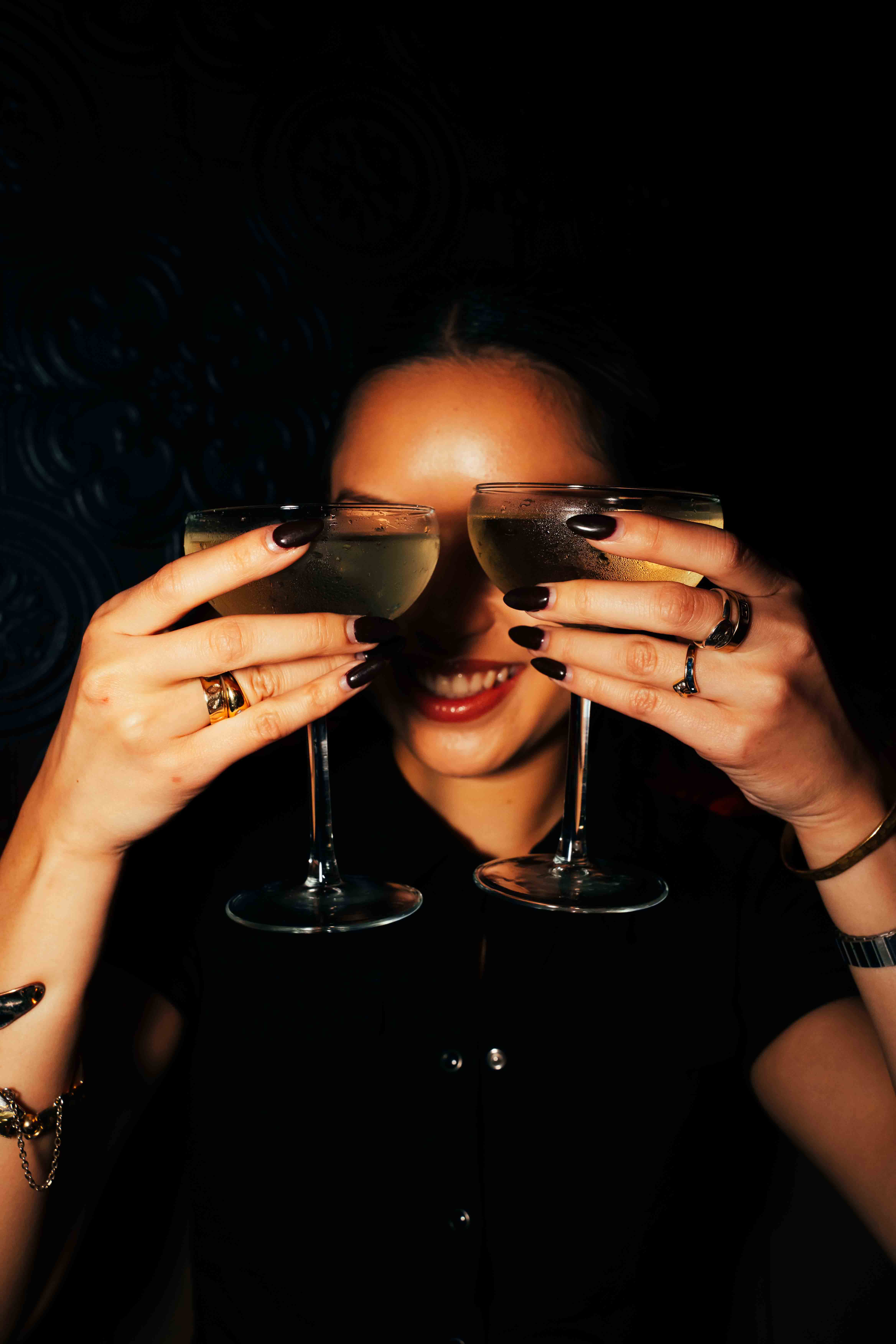 Person with a mustache wearing a black t-shirt carrying a tall stack of pizza boxes inside a dimly lit bar or restaurant.