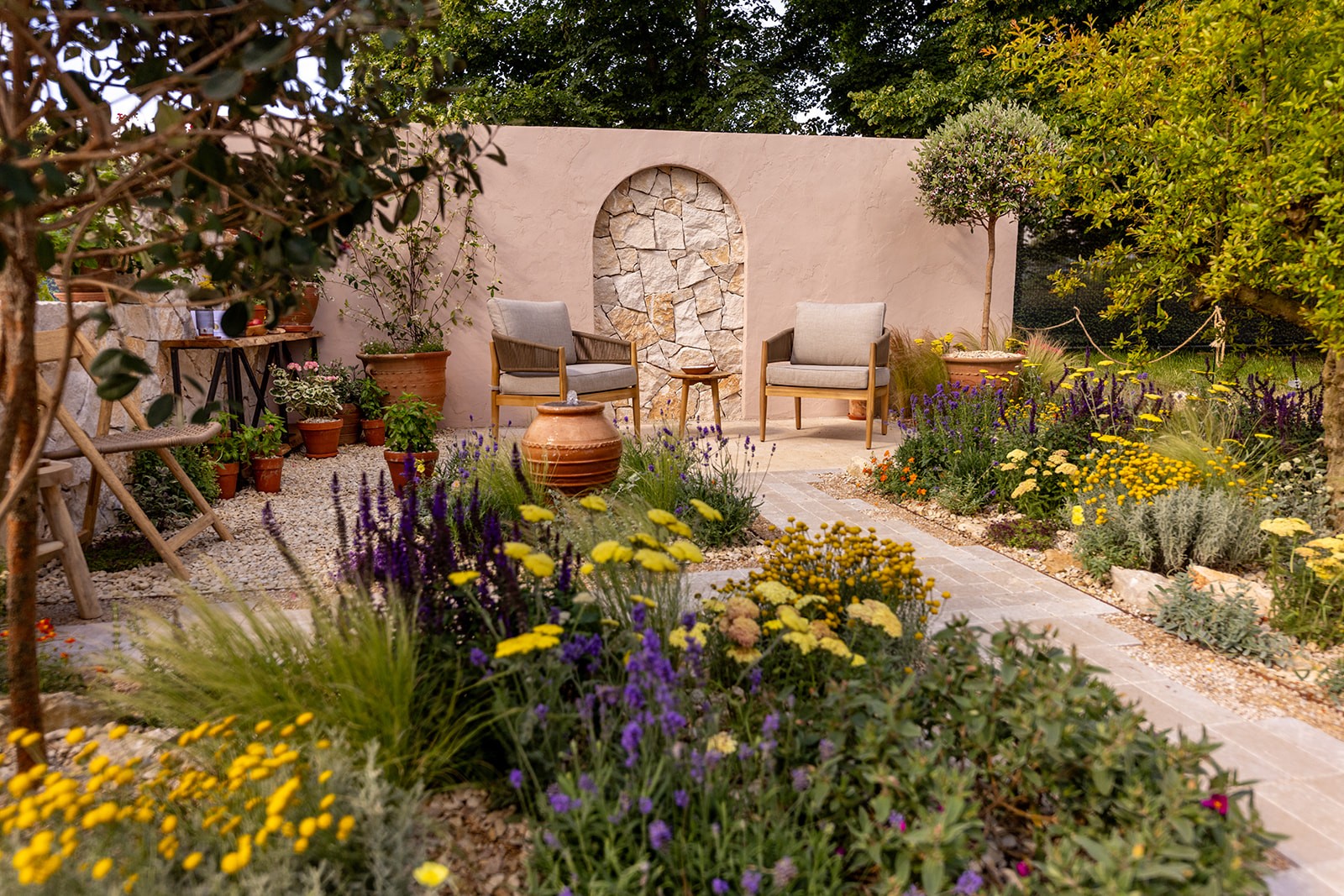 Lush garden with colorful flowers, winding paths, and a stone wall in the background under a sunny sky.