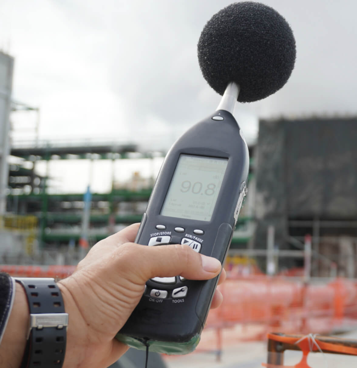 A hand holds a portable sound level meter against a construction site background.