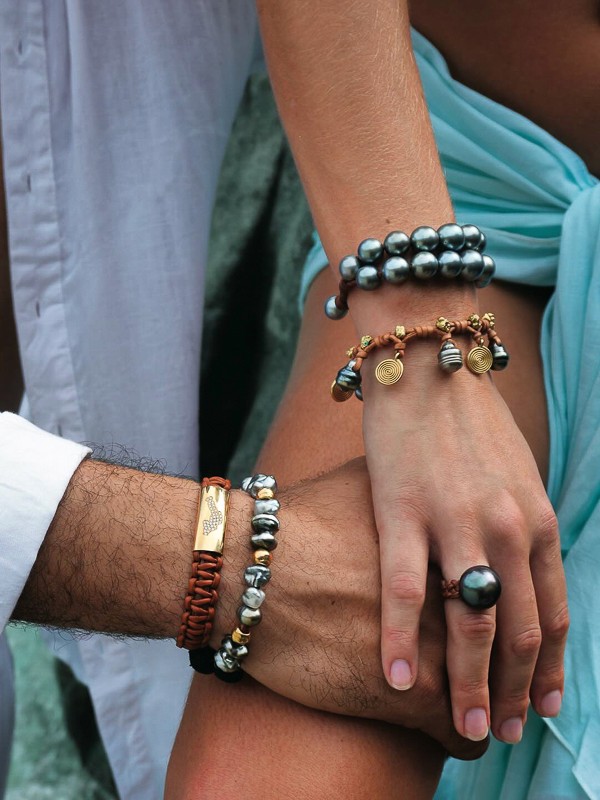 Close-up of intertwined hands adorned with Manos de Oro Saint Barth handcrafted luxury jewelry—featuring his-and-hers leather and pearl bracelets with gold accents and a bold Tahitian pearl ring, symbolizing soulful connection and island elegance.