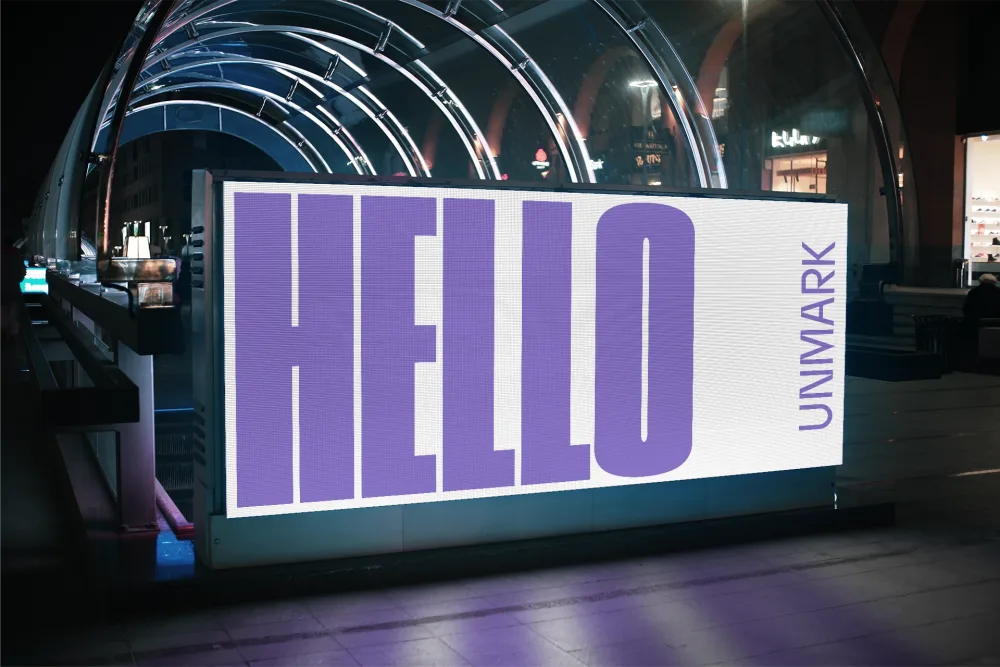 Illuminated outdoor advertising board displaying bold purple text on a white background, positioned under a modern arched glass structure in a city at night.