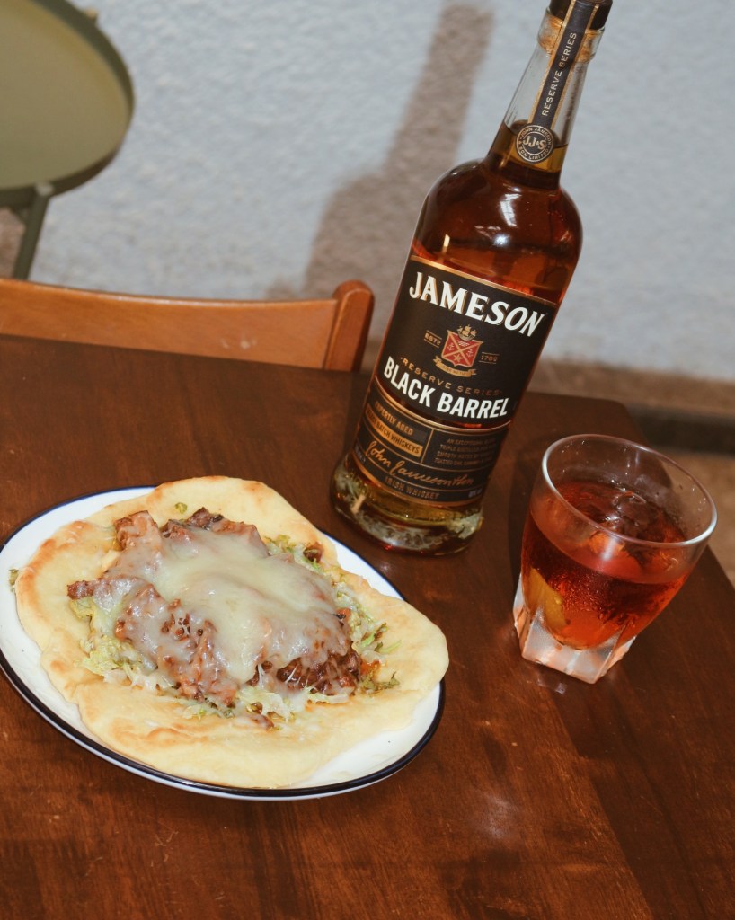 A plate of pulled pork on flatbread topped with melted cheese and lettuce, accompanied by a bottle of Jameson Black Barrel whiskey and a glass of whiskey cocktail on a wooden table.