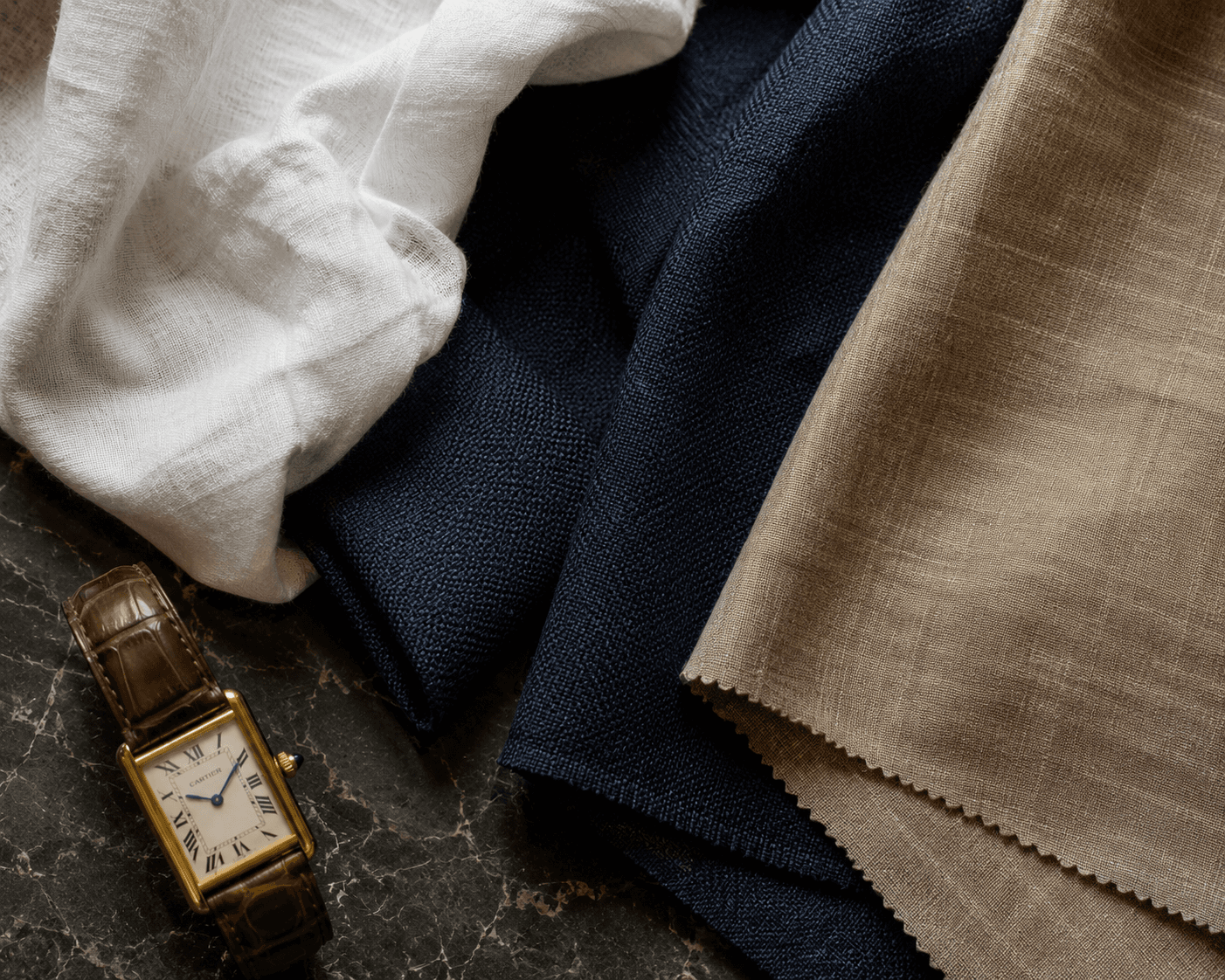 A close-up flat lay of premium menswear textile swatches arranged on a dark marble surface. The arrangement features draped soft white linen, textured deep navy blue hopsack wool, and a swatch of beige linen with pinked zig-zag edges. Resting alongside the fabrics in the lower left corner is a luxury rectangular gold wristwatch with a white dial, black Roman numerals, and a brown alligator leather strap.