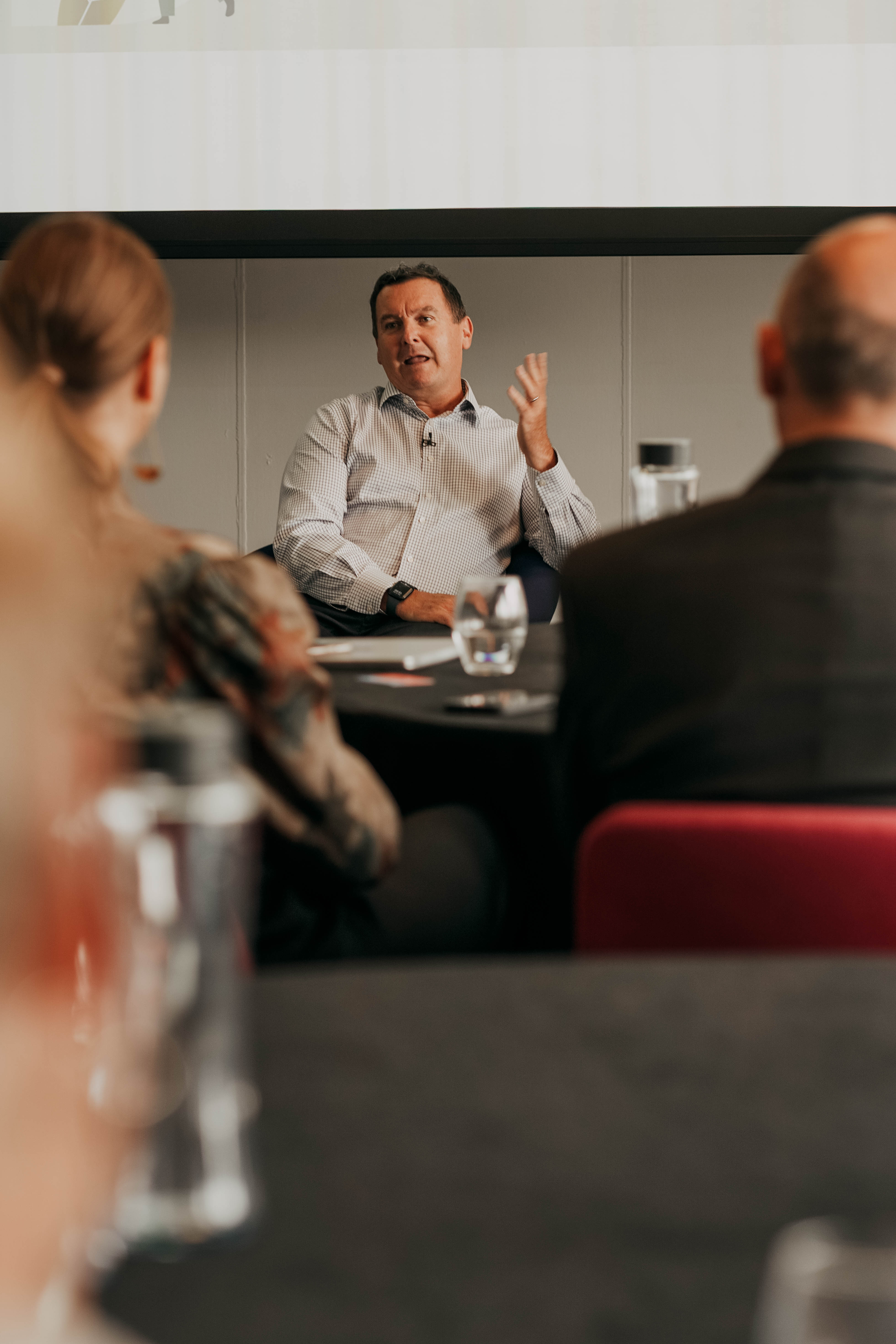 A man in a striped button-down shirt sits at a conference table and speaks expressively, gesturing with one hand, as he addresses other attendees who are seen from behind in the foreground.