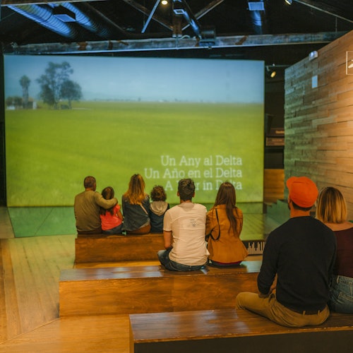 People sitting on wooden steps, watching a projected video of a field with the text "Un Año en el Delta" in multiple languages.