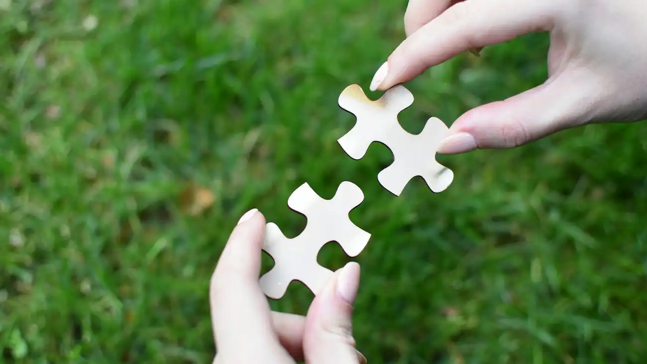 Two hands connecting white puzzle pieces over grass, representing a flexible sponsorship options menu, add-ons and tailored cooperation building blocks for sports clubs.